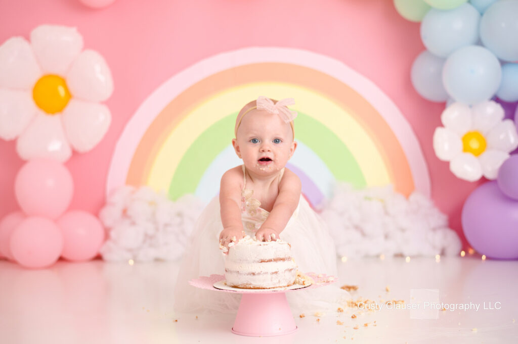 A baby in a white dress and pink bow smiles while grabbing a small frosted cake. The background features a pastel rainbow and large flower-shaped balloons. The scene has a festive and playful atmosphere.