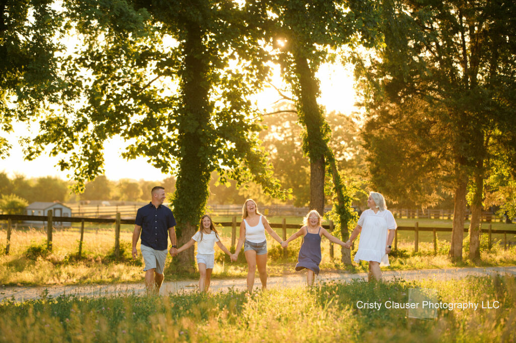 A family of five holds hands and walks together along a sunlit country path, surrounded by green trees and fields, with warm golden light in the background. Cristy Clauser Photography