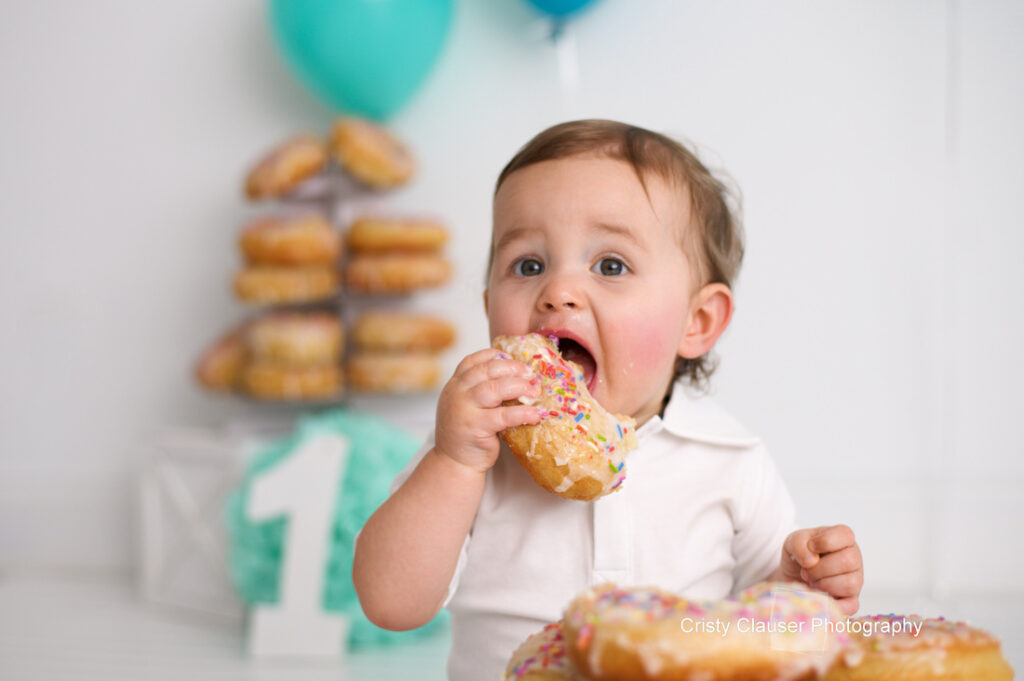 A toddler with light hair and wearing a white shirt is enthusiastically biting into a large donut with colorful sprinkles. Behind the child, there is a stack of donuts, a blue balloon, and a white number one sign.