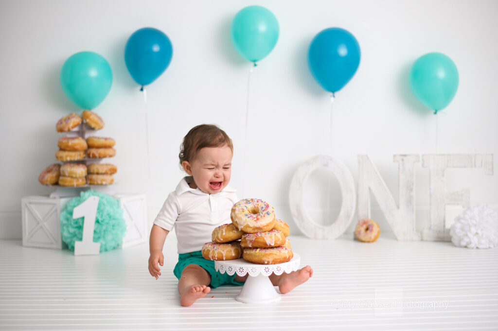 A toddler sits on a white floor surrounded by teal balloons, a stack of donuts, and decorative letters spelling "ONE." The child, wearing a white shirt and teal shorts, is joyfully smiling at donuts placed on a cake stand.