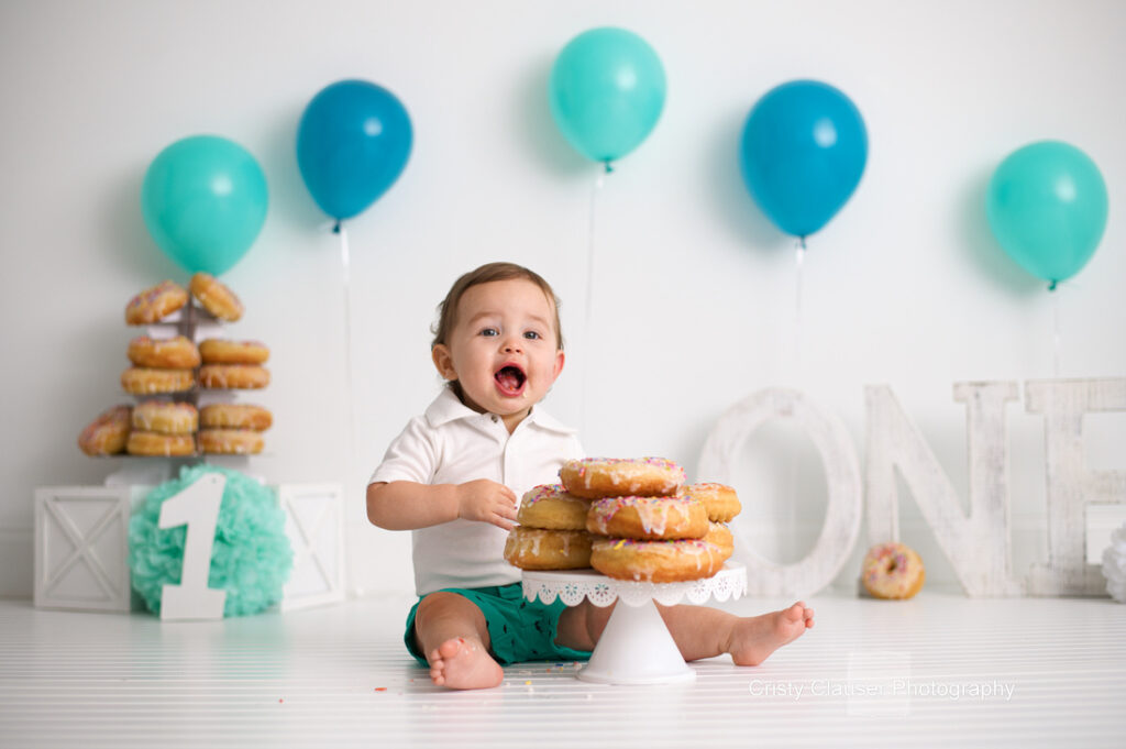 A baby sits on the floor with a cake stand of donuts in front. Behind are teal balloons and stacked donuts. White blocks with a large number "1" and decorative letters spell "ONE" nearby. The baby wears a white shirt and looks cheerful.