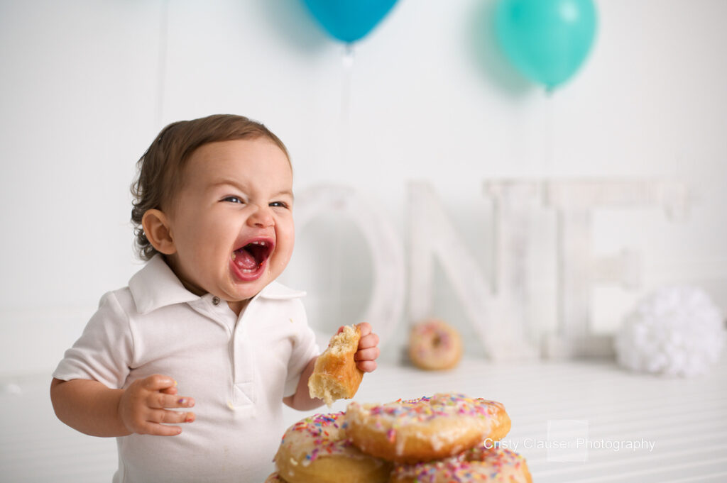 A joyful toddler in a white shirt gleefully holds and eats a donut. In the background, pastel blue balloons and the word "ONE" hint at a first birthday celebration. A stack of sprinkled donuts is in focus.