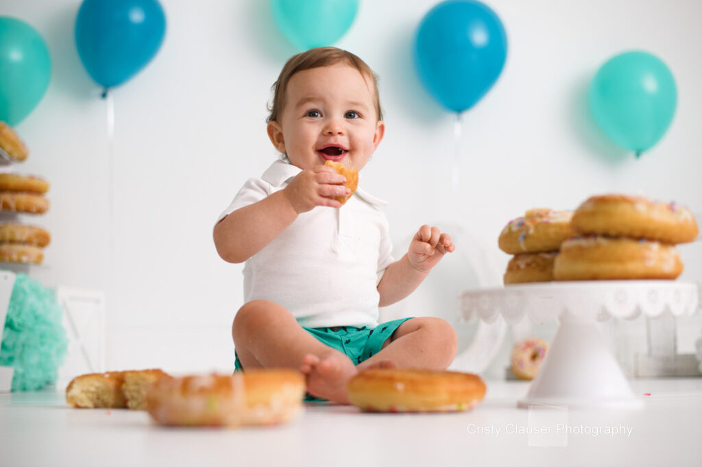 A smiling baby sits on the floor surrounded by donuts, holding one in hand. The baby wears a white shirt and teal shorts. Teal and blue balloons are in the background, suggesting a festive setting.