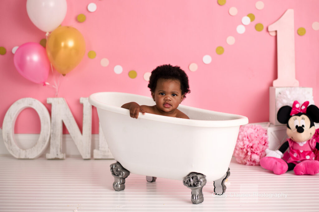 A baby sits in a white clawfoot tub against a pink backdrop. Surrounding the tub are balloons, a large number "1," and a Minnie Mouse stuffed toy. The floor is white and striped.