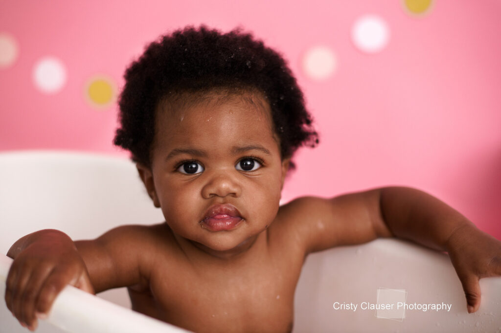 A baby with short curly hair is sitting in a white tub, looking toward the camera. The background is pink with white and yellow dots. The photograph is credited to Cristy Clauser Photography.
