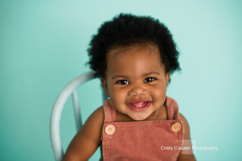 A smiling baby in a brown overall sits on a chair against a light teal background. The baby has curly hair and is looking directly at the camera.