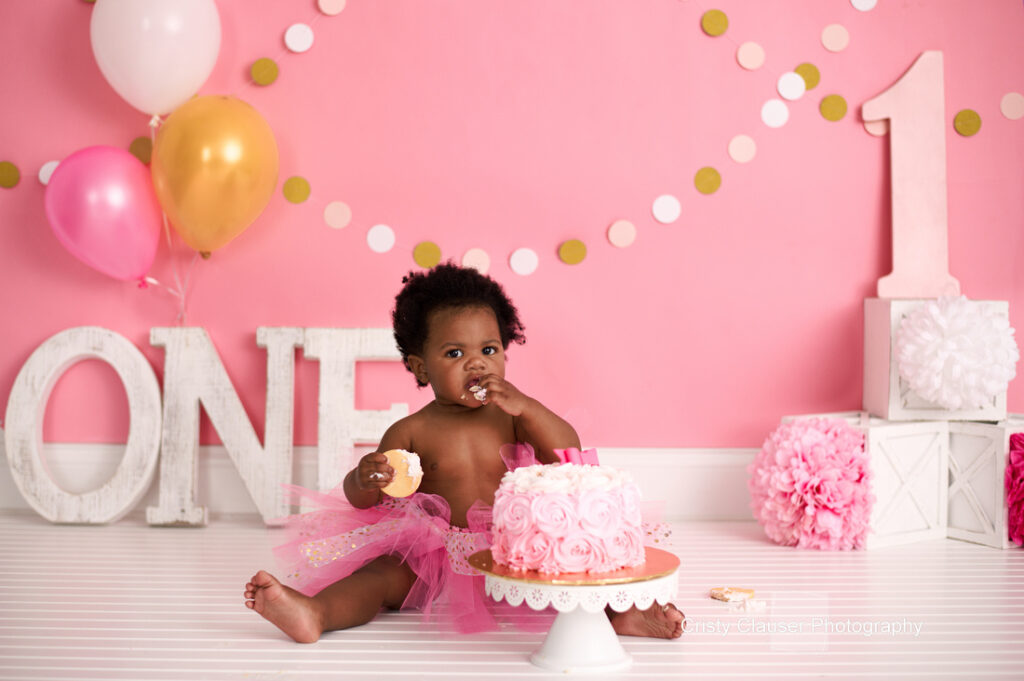 A baby wearing a pink tutu sits on a white floor, eating cake with pink frosting. The background is pink with gold, pink, and white balloons, a large number "1," and the word "ONE" in white letters. Pink and white decorations surround the scene.