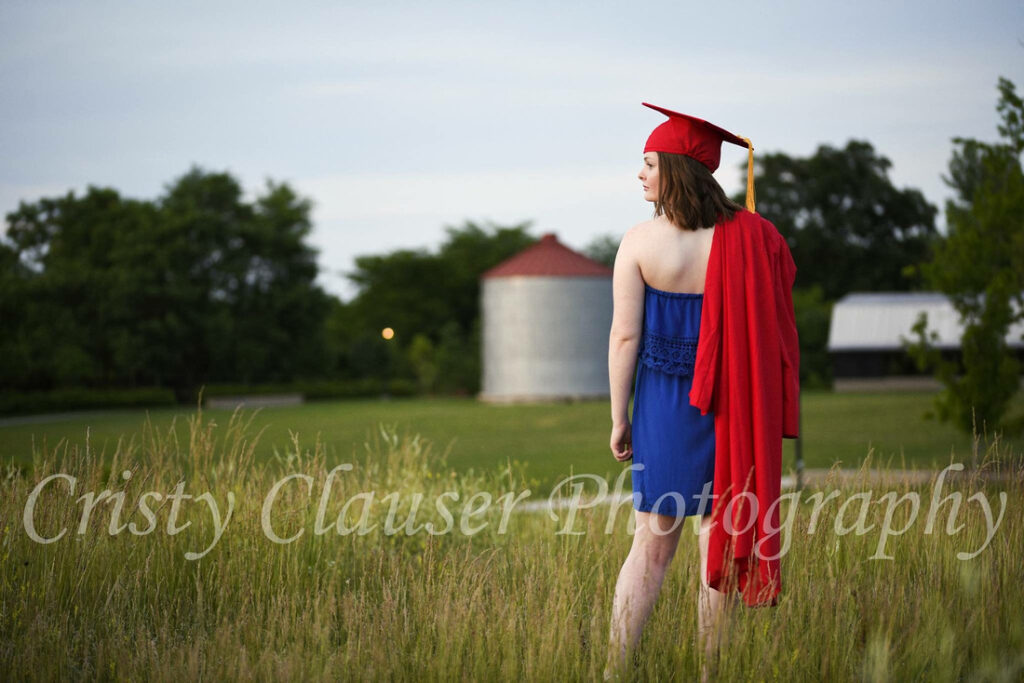 A graduate in a blue dress and red cap and gown stands in a grassy field, holding her gown over her shoulder. Trees, a silo, and a barn are visible in the background. The text "Crsty Clauser Photography" is across the image.