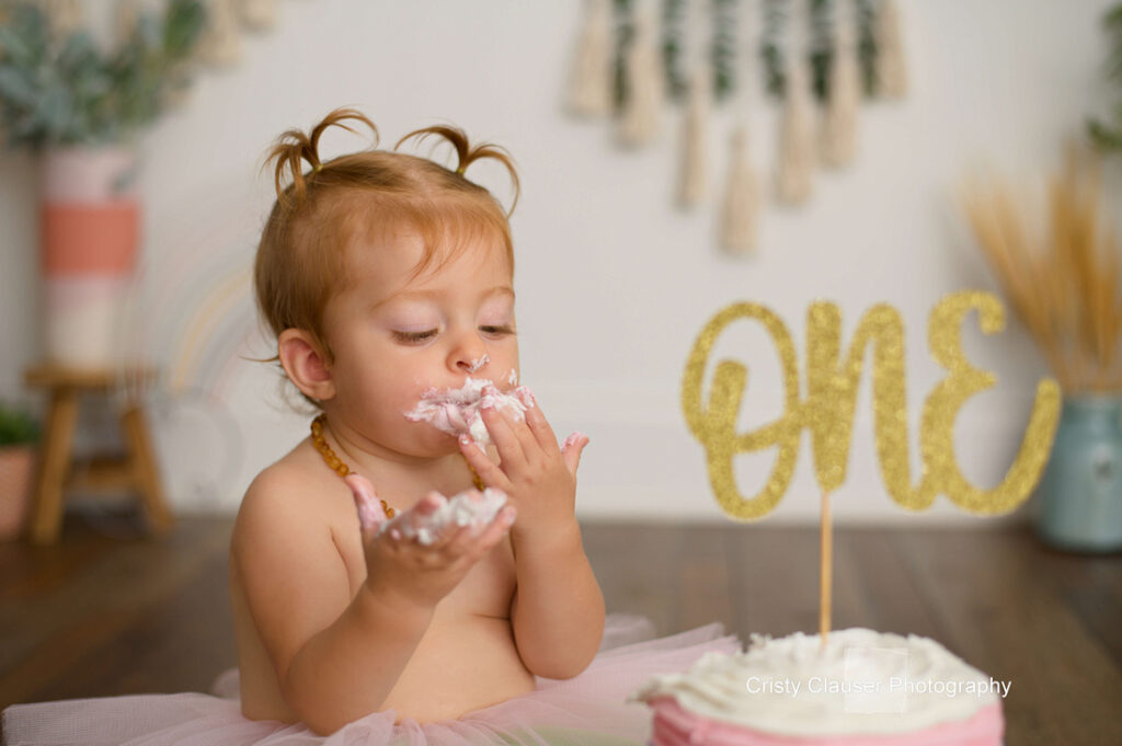 A toddler with light brown hair in pigtails, wearing a pink tutu, eats cake with pink and white frosting. A gold "ONE" sign is on the cake, celebrating her first birthday. The background is softly decorated. Cristy Clauser Photography