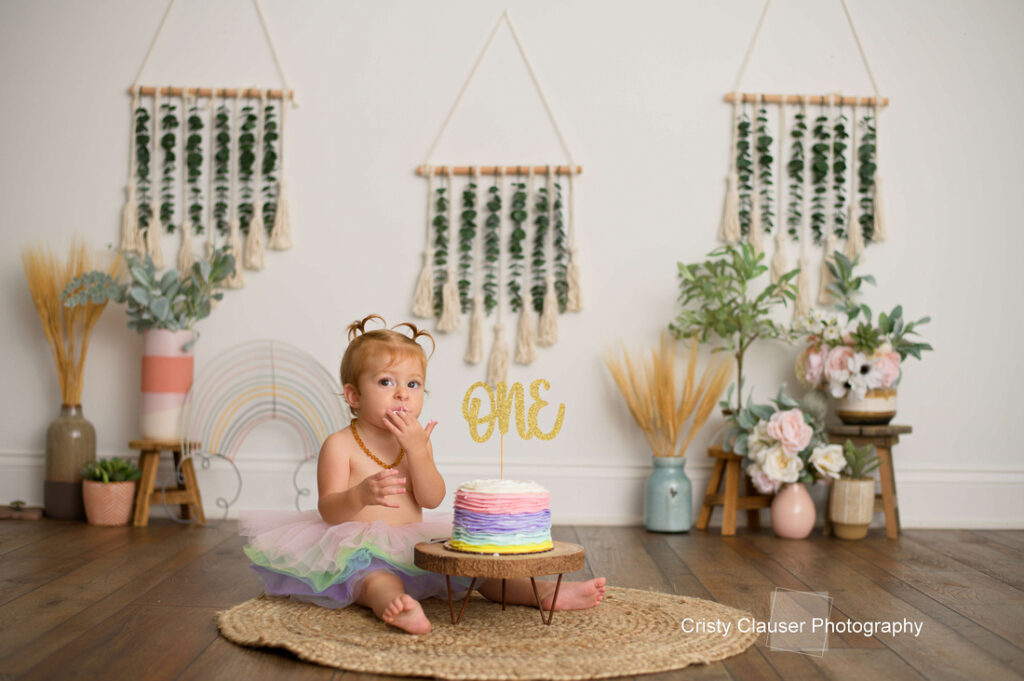 A toddler in a tutu sits on a mat indoors, eating a pastel-colored birthday cake with a gold “ONE” topper. The room is decorated with plants, flowers, and boho wall hangings. Cristy Clauser Photography