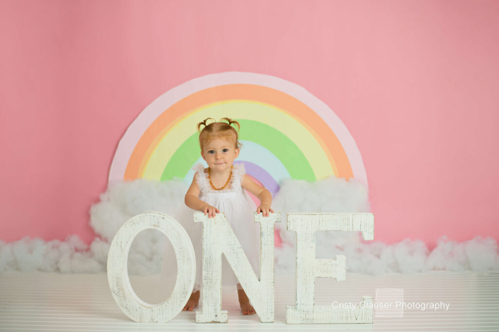 A toddler girl in a white dress stands behind large white letters spelling "ONE," with a pastel rainbow and fluffy clouds against a pink background, celebrating her first birthday. Cristy Clauser Photography