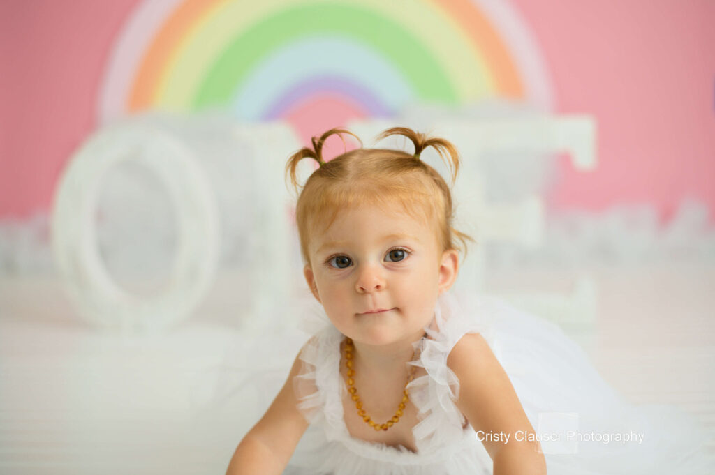 A young child with light brown hair in pigtails wears a white dress and amber necklace, crawling on a white floor. A pastel rainbow and large white "ONE" letters are in the blurred background. Cristy Clauser Photography