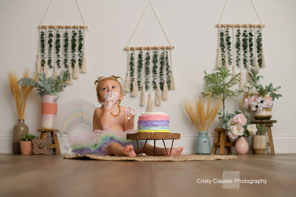 A baby wearing a colorful tutu sits on a rug, eating a pastel rainbow cake with their hands. The backdrop includes macrame wall hangings, flowers, and greenery in vases, creating a boho-style setting. Cristy Clauser Photography