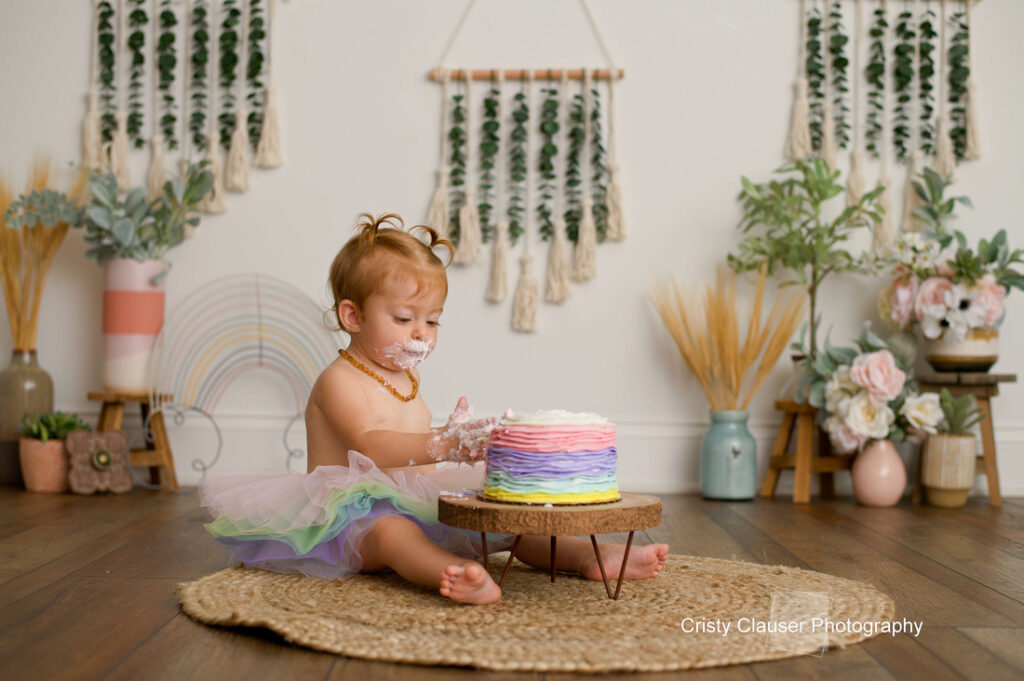 A toddler in a pastel tutu sits on a rug, happily eating a rainbow-layered cake. The room is decorated with plants, flowers, and woven wall hangings, creating a whimsical, boho atmosphere. Cristy Clauser Photography