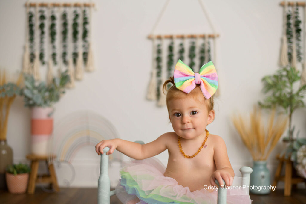 A toddler wearing a large pastel rainbow bow and beaded necklace sits on a chair, surrounded by soft-colored plants and boho decor. She has a slight smile and is dressed in a pastel tulle skirt. Cristy Clauser Photography