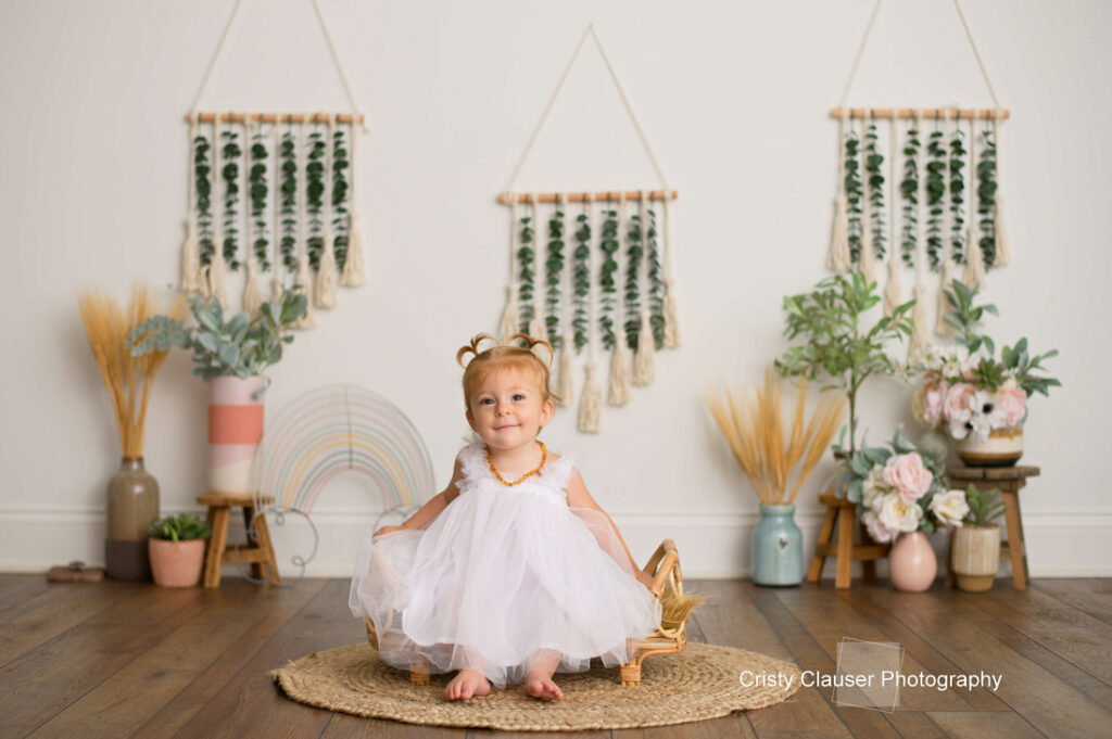 A young toddler in a white dress sits barefoot on a round rug, smiling. She is surrounded by plants, flowers, and boho decor, with three green wall hangings behind her. The scene is bright and cheerful. Cristy Clauser Photography