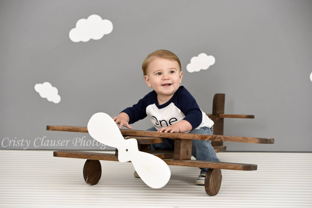 A smiling toddler sits in a wooden toy airplane against a gray background with white clouds. He wears a navy blue and white shirt with "one" on it, celebrating his first birthday. Cristy Clauser Photography