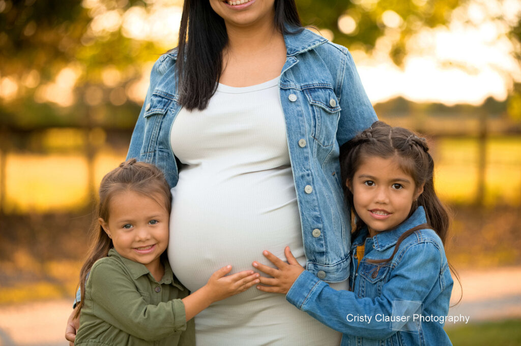 A pregnant woman in a denim jacket stands outdoors, smiling, with two young girls hugging her and touching her belly. The girls look happy, and the background is softly blurred with warm sunlight. Cristy Clauser Photography