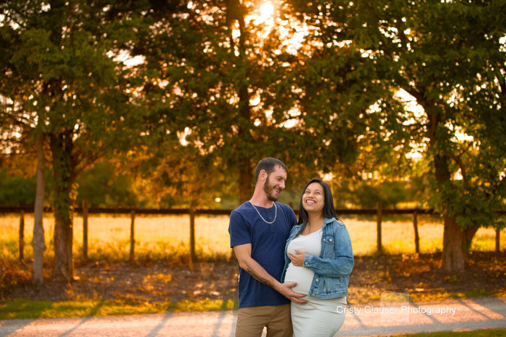 A smiling couple stands outdoors at sunset; the man gently touches the woman’s pregnant belly as she holds his hand. Trees and golden sunlight create a warm, serene background. Cristy Clauser Photography