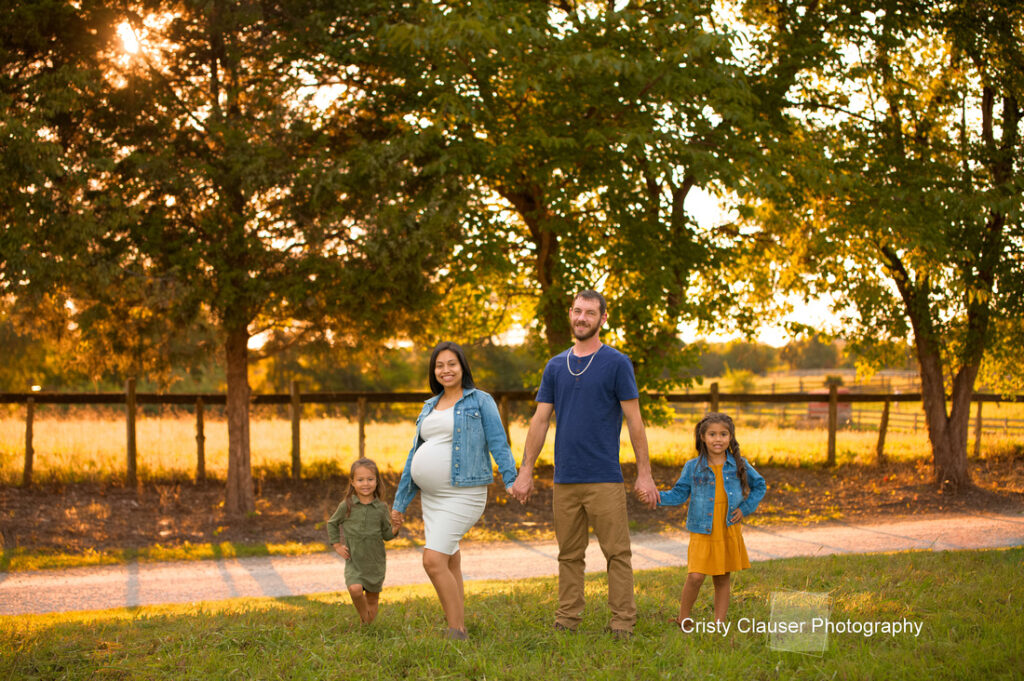 A pregnant woman, a man, and two young children stand holding hands in a grassy field with trees and sunlight in the background. The family looks happy and relaxed, enjoying a warm day outdoors. Cristy Clauser Photography