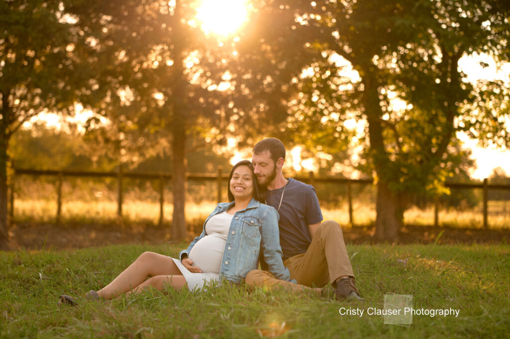 A smiling pregnant woman sits on the grass, leaning against a man as they enjoy a sunny outdoor setting with trees and a wooden fence in the background. The photo is warmly lit by the setting sun. Cristy Clauser Photography