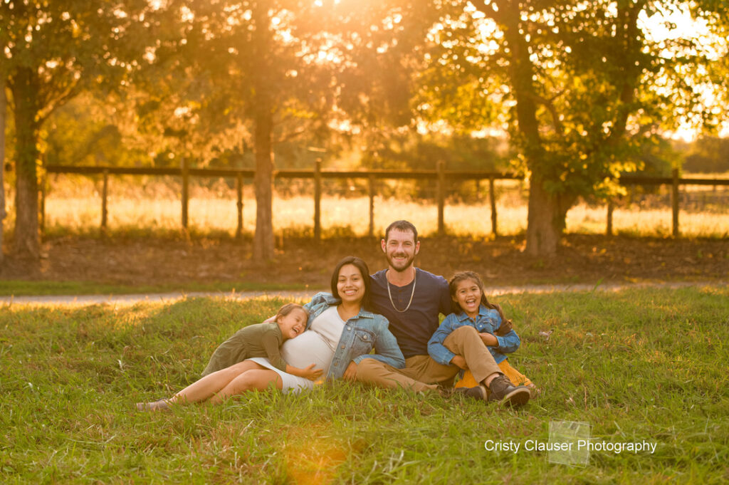 A smiling family of four sits on grass at sunset. The pregnant mother sits in the center, with her partner and two young children leaning happily against her. Sunlight filters through trees in the background. Cristy Clauser Photography