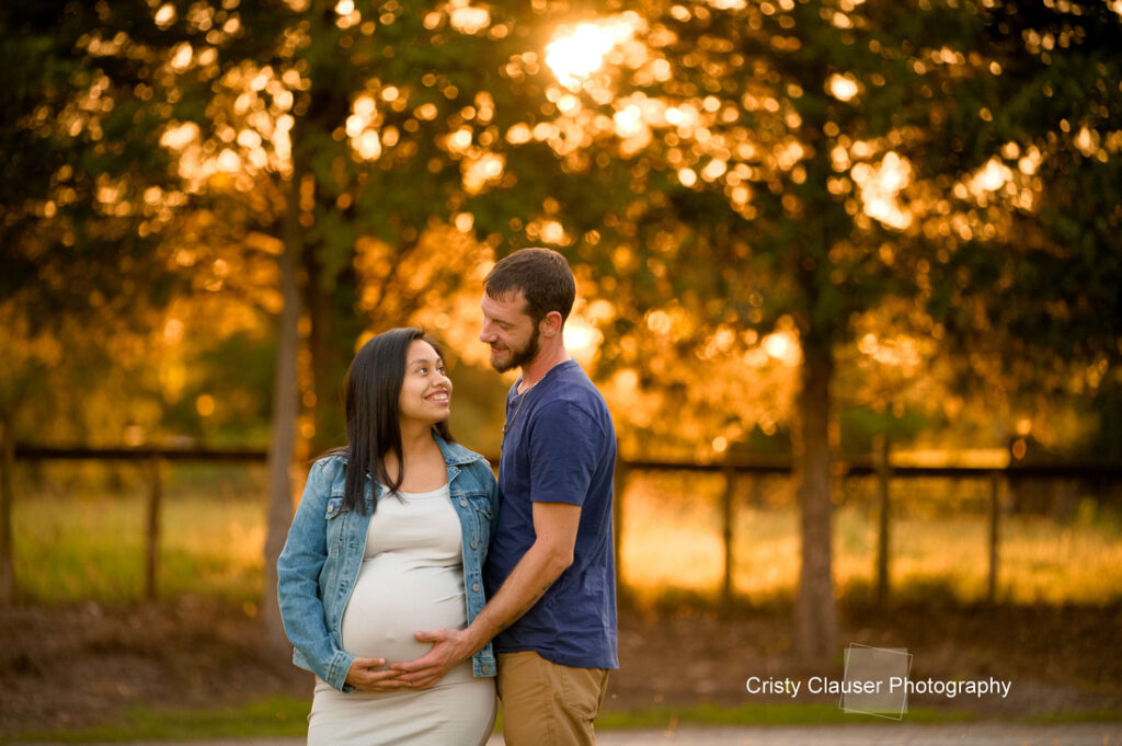 A smiling couple stands outdoors at sunset; the man gently holds the woman's pregnant belly as they gaze at each other, surrounded by trees and warm golden light. Cristy Clauser Photography