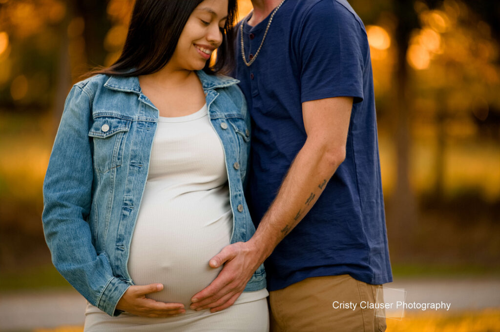 A pregnant woman in a white dress and denim jacket smiles as she and her partner gently cradle her baby bump together outdoors at sunset. Cristy Clauser Photography