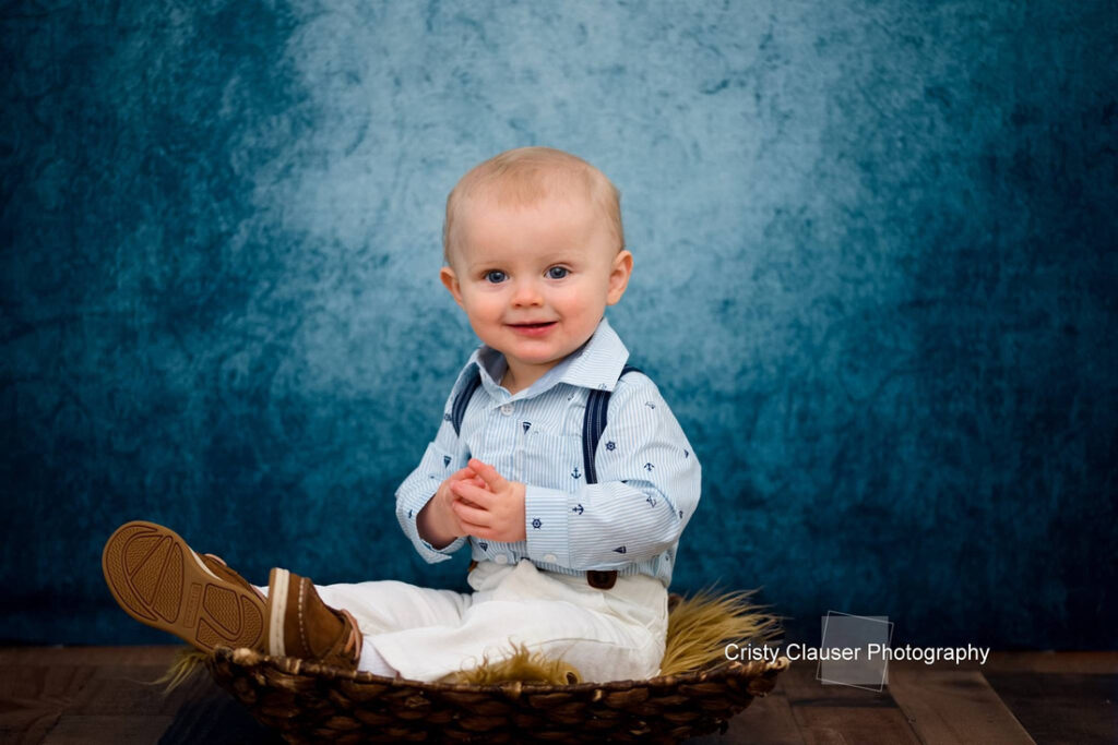 A smiling toddler with short blond hair sits in a woven basket on a wooden floor, wearing a light blue shirt, suspenders, and white pants, in front of a blue textured background. Cristy Clauser Photography