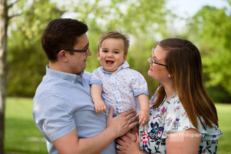A smiling baby is held between two adults outdoors, all looking at each other lovingly on a sunny day with green trees in the background. The adults wear glasses and light-colored shirts. Cristy Clauser Photography