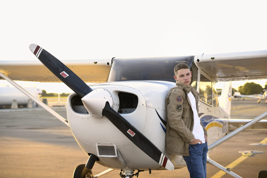 A person in a jacket and scarf leans against the nose of a small airplane parked on a runway at sunset. The propeller is visible, and the sky is clear, casting a warm light on the scene.