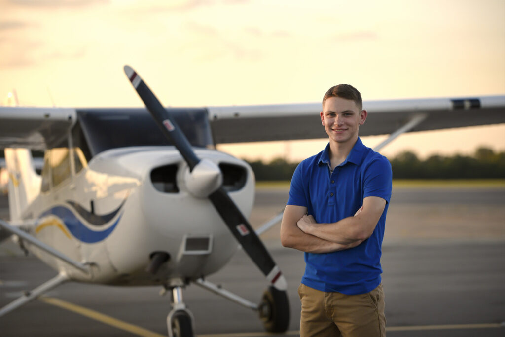 A young person in a blue polo shirt stands with arms crossed in front of a small propeller airplane on a runway at sunset. The background shows a clear sky and a tree line.