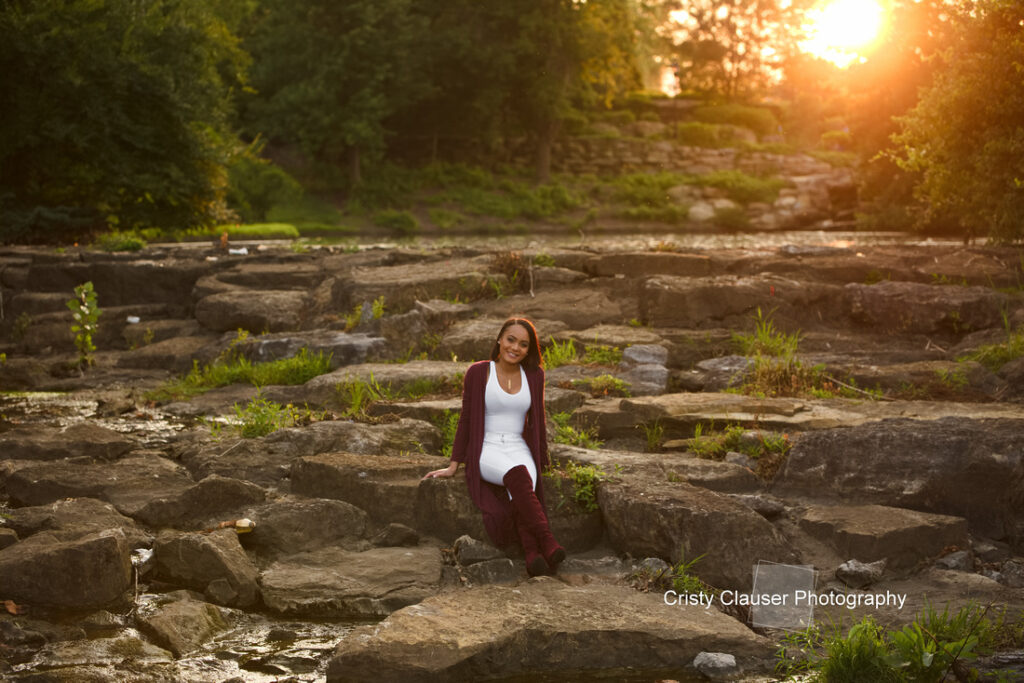 Woman sitting on large rocks with a serene expression. She's wearing a white outfit and a maroon cardigan, surrounded by lush greenery. The sun sets in the background, casting a warm glow. The image is signed "Cristy Clauser Photography.