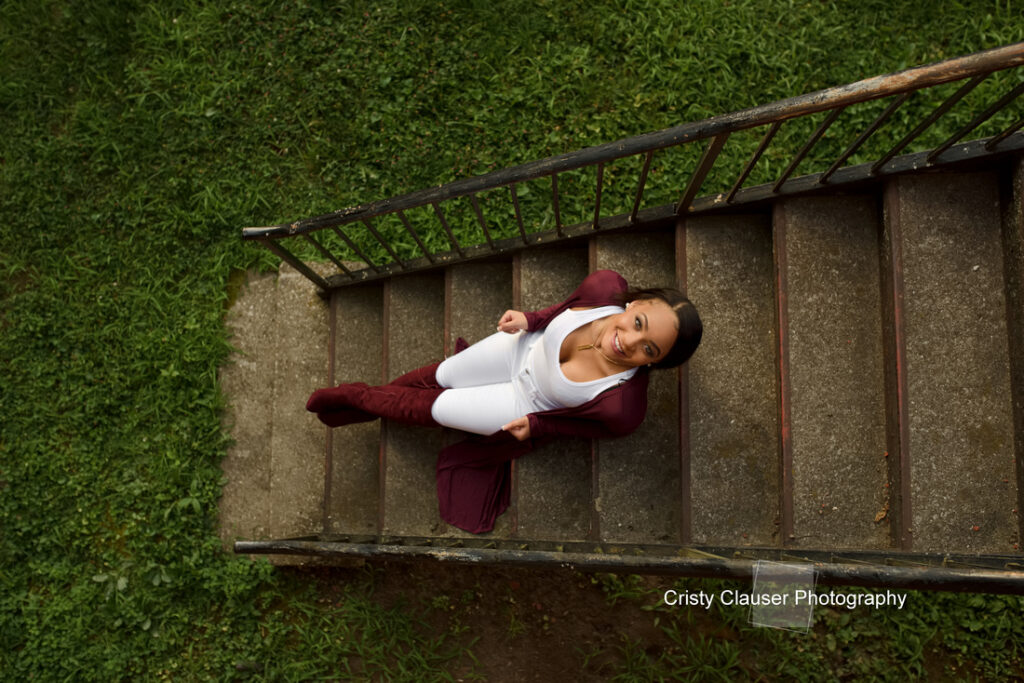 A person in a maroon jacket and white pants stands on an outdoor staircase, looking up with a smile. Lush green grass surrounds the staircase. The photo is credited to Cristy Clauser Photography.