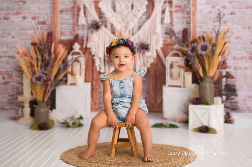 A toddler in a blue romper and floral headband sits smiling on a small wooden stool. The background features rustic decor, dried flowers, and macramé against a white brick wall. Cristy Clauser Photography