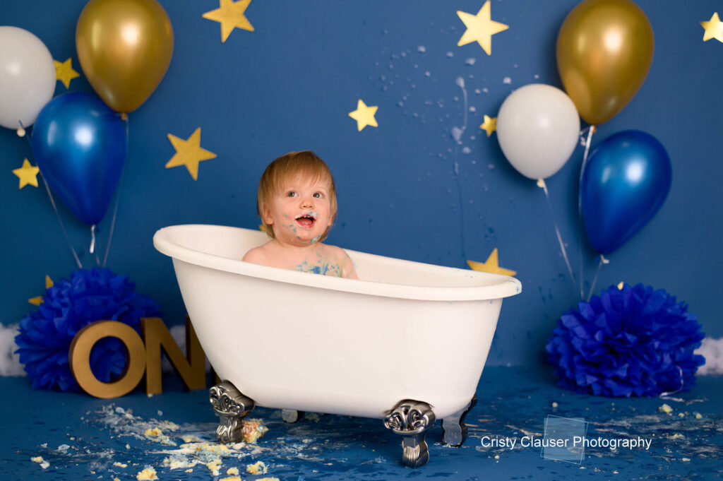 A smiling baby with cake on their face sits in a white clawfoot bathtub. The background is blue with gold stars, blue and gold balloons, and blue decorations. The floor is messy with bits of cake. Cristy Clauser Photography