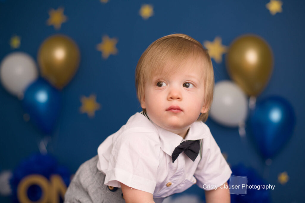 A young boy in a white shirt and black bow tie looks to the side with a serious expression. The background features blue and gold balloons and star decorations, suggesting a festive or birthday setting. Cristy Clauser Photography