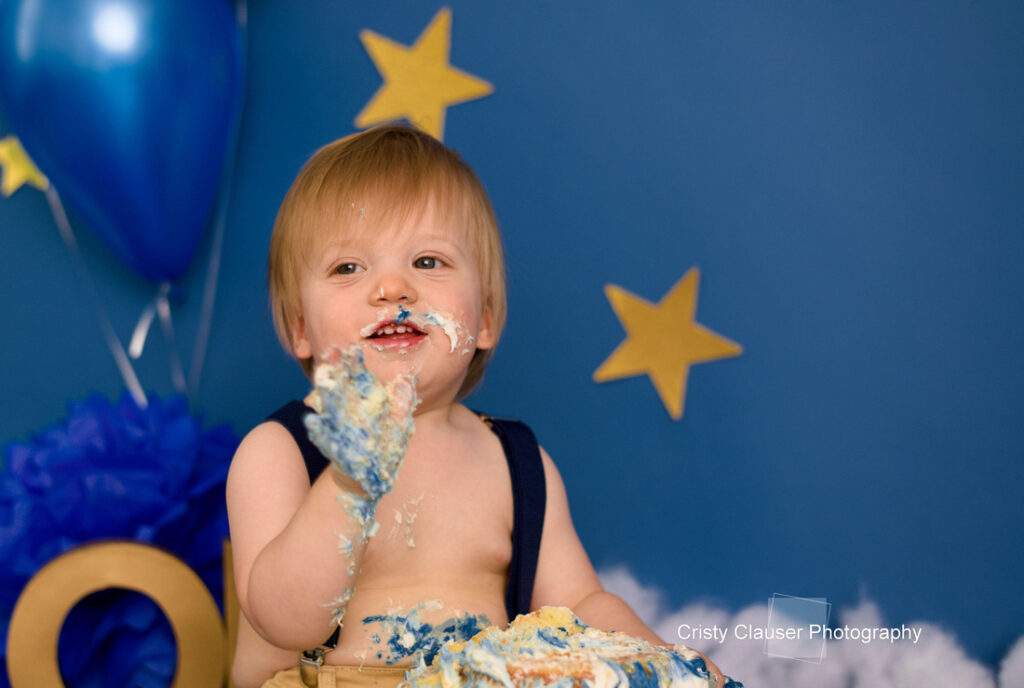 A toddler with blonde hair enjoys a messy cake smash, with blue and yellow frosting on his face and hands. He sits in front of a blue backdrop decorated with yellow stars and balloons. Cristy Clauser Photography