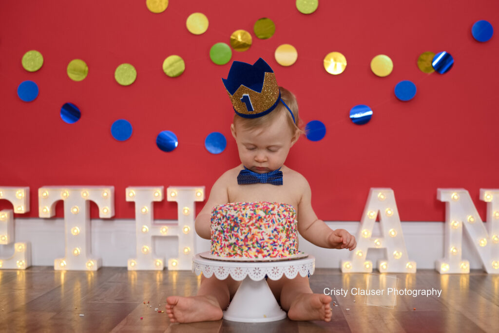A baby wearing a blue crown and bow tie sits in front of a sprinkle-covered cake, with illuminated letters spelling “ETHAN” and colorful decorations in the background. Cristy Clauser Photography
