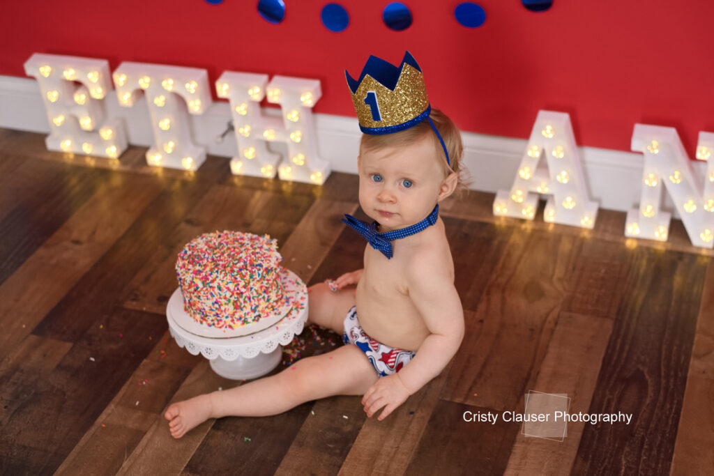 A baby in a blue crown and bow tie sits on a wooden floor next to a sprinkle-covered cake. Large, lit-up letters spell out a name in the background. The photo is by Cristy Clauser Photography. Cristy Clauser Photography