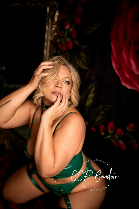 A woman with blonde hair poses confidently in green lingerie against a dark, floral background. She gazes upward, touching her face with one hand and her hair with the other. "CCP Boudoir Photography LLC" watermark is visible. Cristy Clauser Photography