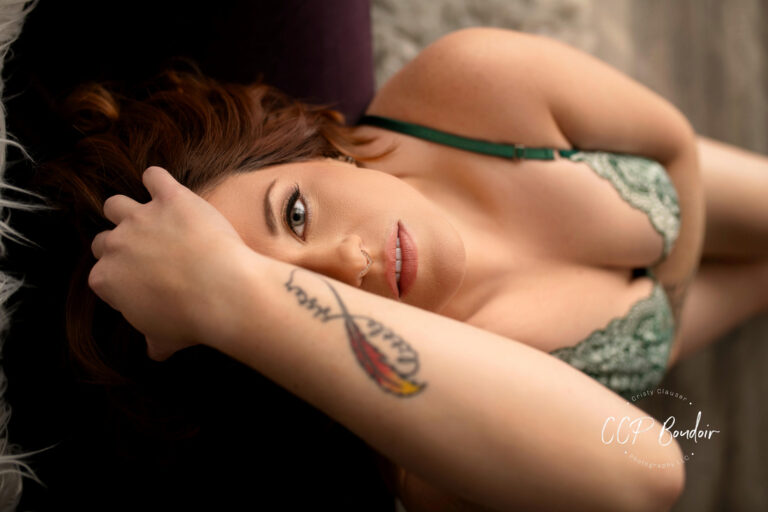 A woman with long hair and a feather tattoo on her forearm lies on her back, wearing green lace lingerie, looking up at the camera with a relaxed expression. The photo has soft lighting and a sensual tone. Cristy Clauser Photography