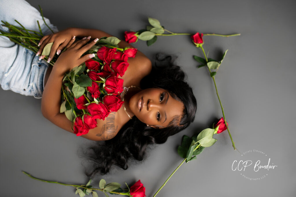 A woman with long dark hair lies on a gray surface holding a bouquet of red roses, with more roses scattered around her. She gazes upwards, wearing light blue jeans and a strapless top. Cristy Clauser Photography