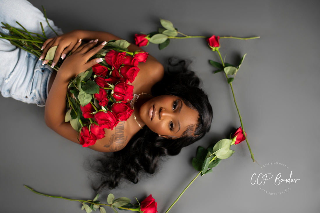 A woman with long dark hair lies on a gray surface holding a bouquet of red roses, with more roses scattered around her. She gazes upwards, wearing light blue jeans and a strapless top. Cristy Clauser Photography