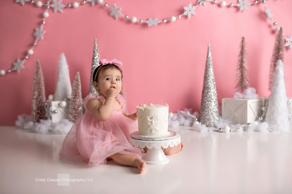 A baby in a pink dress sits on the floor in front of a white cake, with one finger in her mouth. The background features pink and white Christmas trees, snowflakes, and wrapped gifts against a pink wall. Cristy Clauser Photography