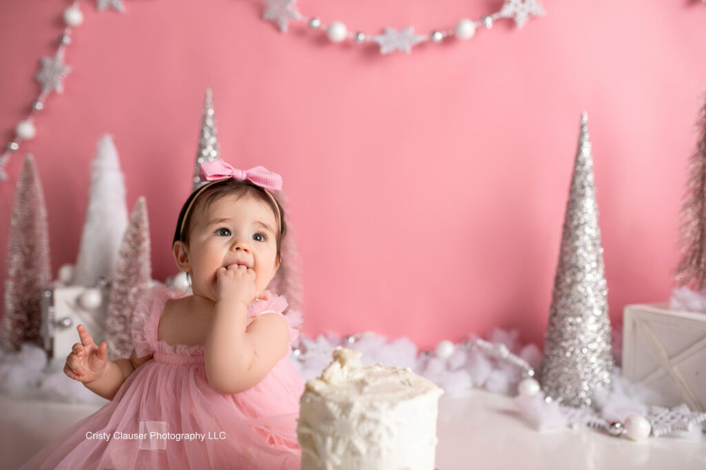 A baby in a pink dress and bow sits in front of a white cake, with silver and white Christmas trees and star decorations on a pink background. The baby has one hand in her mouth and is looking up. Cristy Clauser Photography