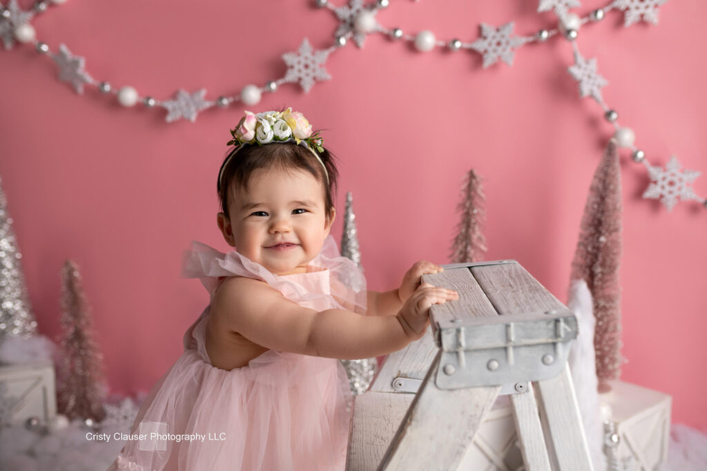 A smiling baby in a pink dress and flower crown stands by a white step ladder. The background is pink with white snowflakes, sparkly trees, and festive decorations. Cristy Clauser Photography