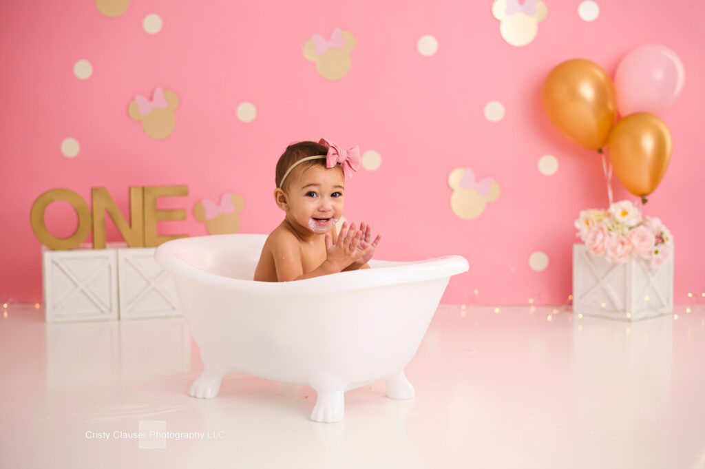 A smiling baby with a pink bow sits in a white clawfoot bathtub. The background is pink with gold polka dots and mouse ears, gold balloons, flowers, and a gold "ONE" sign, celebrating a first birthday. Cristy Clauser Photography