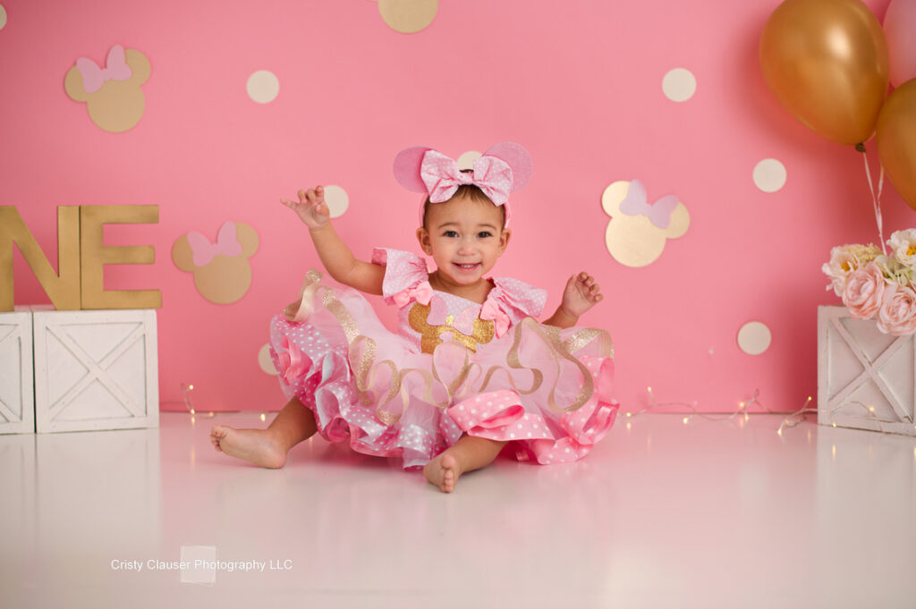 A smiling toddler in a pink ruffled Minnie Mouse dress and headband sits on the floor with arms raised. The background is pink with gold Minnie Mouse shapes, balloons, white crates, and flowers. Cristy Clauser Photography