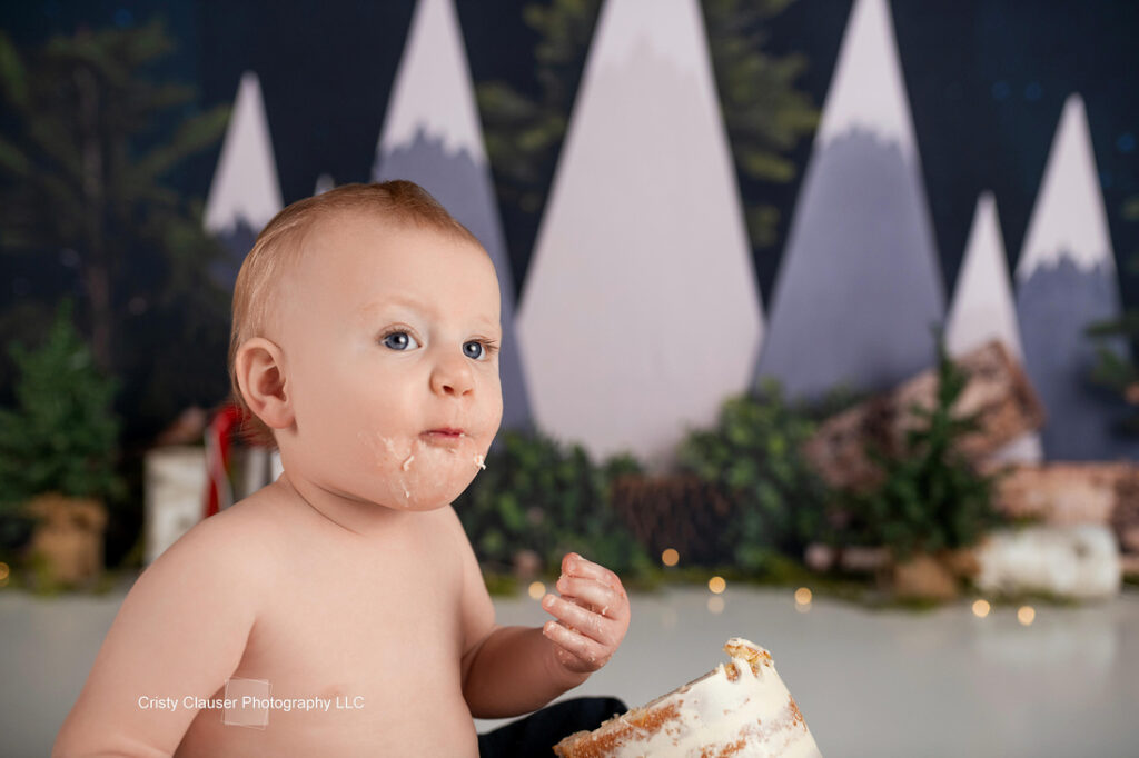 A baby with frosting on their face and hands sits shirtless in front of a cake, with a whimsical winter forest and mountain backdrop behind them. Cristy Clauser Photography